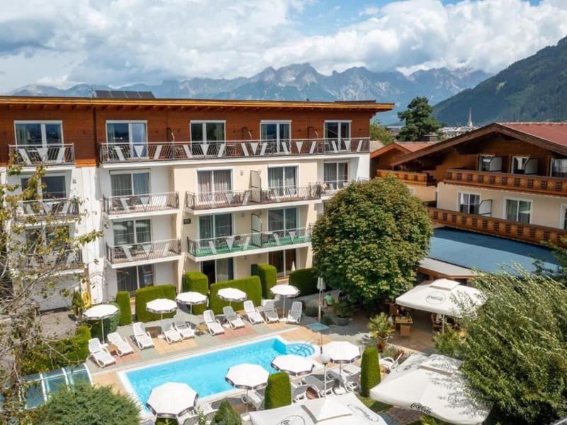 Hotelgebäude mit Swimmingpool, Liegestühlen, Bäumen und Bergen im Hintergrund unter bewölktem Himmel.