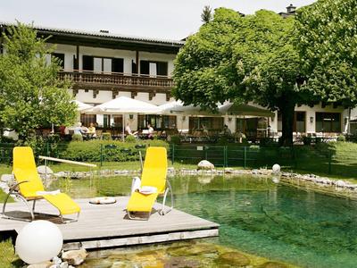 Garten mit großem Teich, Holzsteg, zwei gelben Liegestühlen und Baum vor einem zweistöckigen Haus.