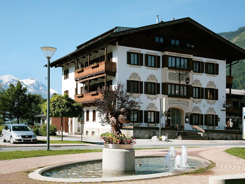 Dreistöckiges weißes Haus mit Holzbalkonen, davor runder Brunnen und Park mit Bergen im Hintergrund.