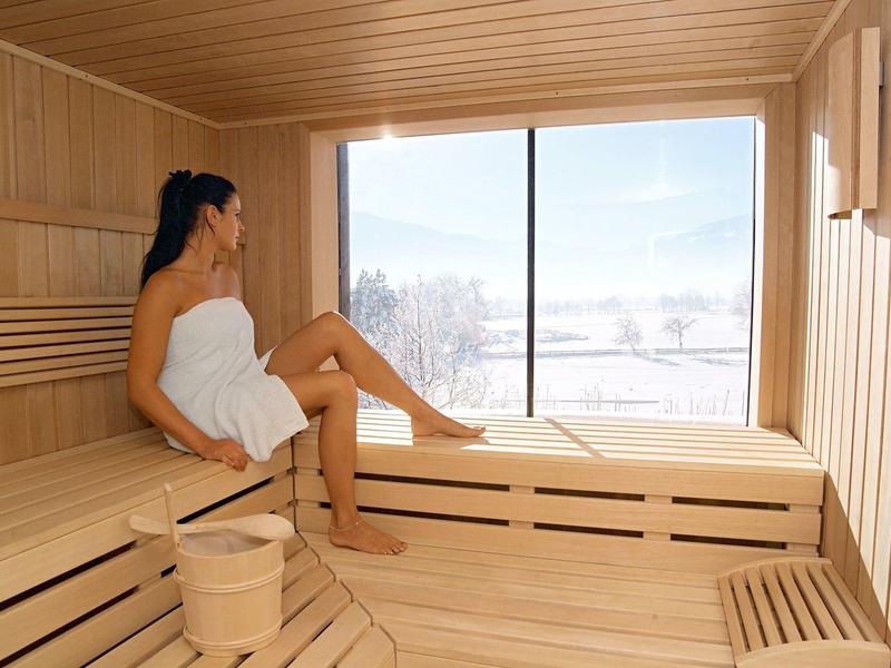 Frau mit Handtuch sitzt in heller Holzsauna vor großem Fenster mit Winterlandschaft draußen.