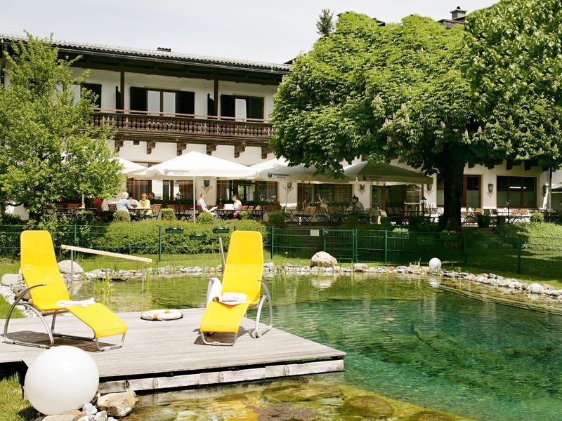 Garten mit großem Teich, Holzsteg, zwei gelben Liegestühlen und Baum vor einem zweistöckigen Haus.