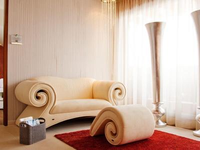 Helles Wohnzimmer mit beiger, geschwungener Couch, passendem Hocker und rotem Teppich vor großen Fenstern.