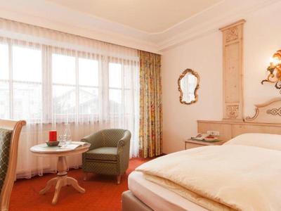 Helles Hotelzimmer mit Einzelbett, grünem Sessel, Vorhängen und großem Fenster mit Tageslicht.