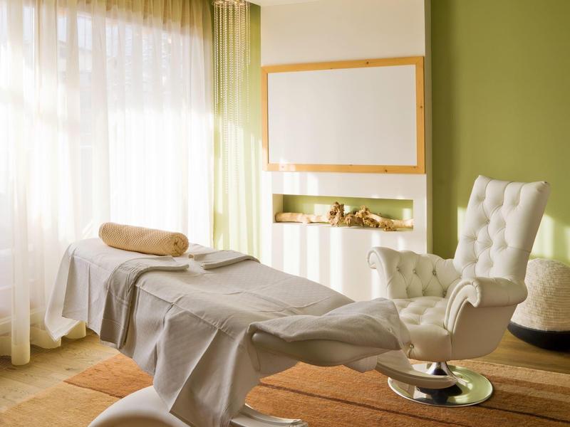Helles Massagestudio mit weißer Liege, beigem Handtuch und gepolstertem Stuhl vor grüner Wand.