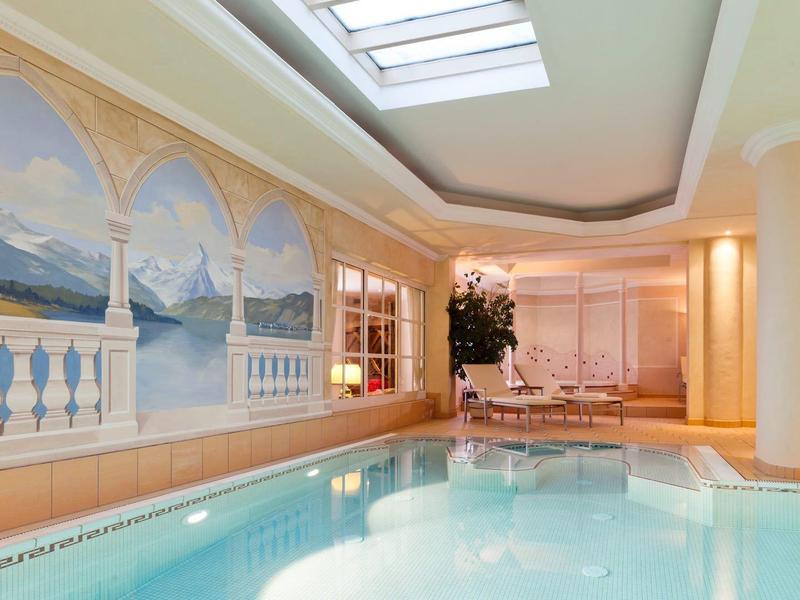 Innenraum mit hellem Hallenbad, blauer Wasserfläche und Wandmalerei mit Landschaftsmotiven.