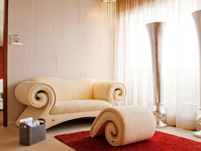 Helles Wohnzimmer mit beiger, geschwungener Couch, passendem Hocker und rotem Teppich vor großen Fenstern.