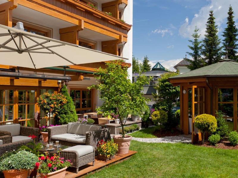 Garten mit Terrasse, Holzgartenhaus, grünen Pflanzen, grauen Möbeln und großem Sonnenschirm.