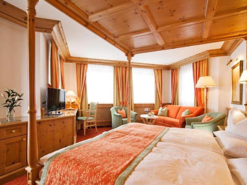 Gemütliches Hotelzimmer mit Holzdecke, Doppelbett, orangefarbenen Vorhängen und Sitzecke.
