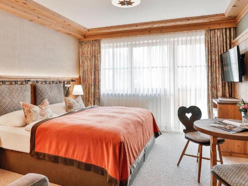 Gemütliches Hotelzimmer mit Doppelbett, Holzmöbeln, orangefarbenem Bettüberwurf und großen Fenstern.