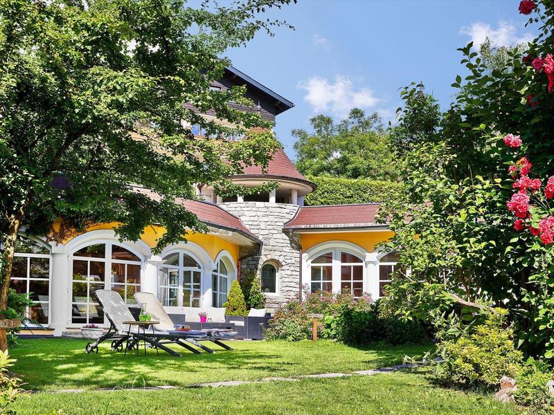 Wohnhaus mit Rundbogenfenstern, Terrasse, Garten, Rasen und blühenden Büschen bei Sonnenschein.