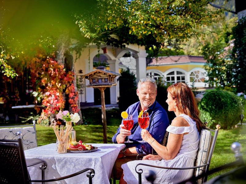 Ein älteres Paar sitzt im Garten und stößt mit Rotweingläsern an, umgeben von grünem Laub.