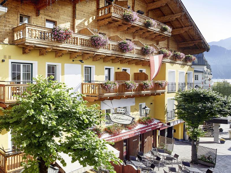 Mehrstöckiges gelbes Alpenhaus mit Holzbalkonen und blühenden Pflanzen an sonnigem Tag.