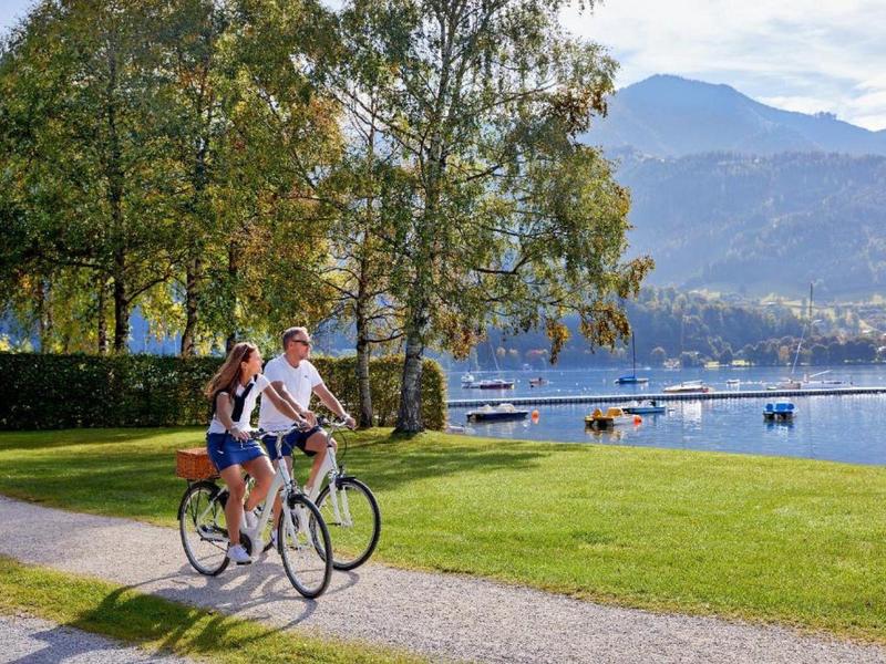 Zwei Personen fahren bei sonnigem Wetter auf einem Weg am Seeufer mit Bergblick Fahrrad.