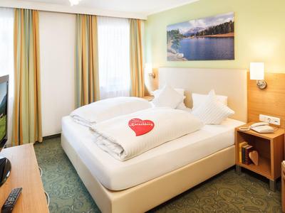 Hell eingerichtetes Hotelzimmer mit Doppelbett, grünem Teppich und Landschaftsbild an der Wand.