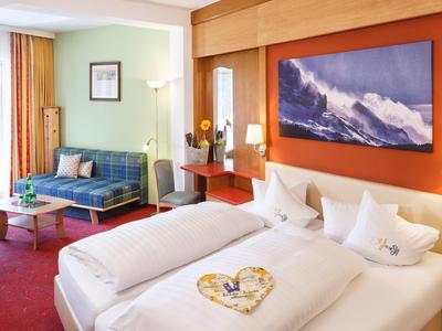 Gemütliches Hotelzimmer mit Doppelbett, Sofa, großem Bergbild und rotem Teppichboden.