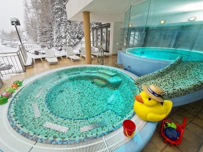 Innenbereich mit rundem Whirlpool mit Mosaikfliesen, Spielsachen und Glaswand, dahinter Schneelandschaft.