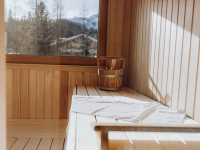 Holzsauna mit Fenster, Holzbänken und Holzeimer, durch das Fenster Blick auf Bäume und Berge.