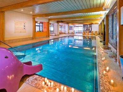 Innenraum mit einem großen Schwimmbad, Holzwänden, Glasfenstern und einem pinken Kinder-Wasserrutsche.