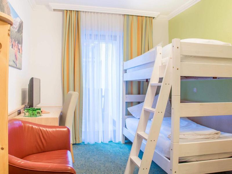 Helles Kinderzimmer mit Etagenbett, kleinem Fernseher, rotem Sessel und Balkon mit Gardinen.