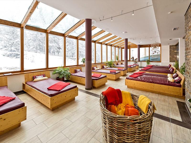 Großer Ruheraum mit Holzbänken, bunten Kissen und bodentiefen Fenstern mit Blick ins Schneelandschaft.