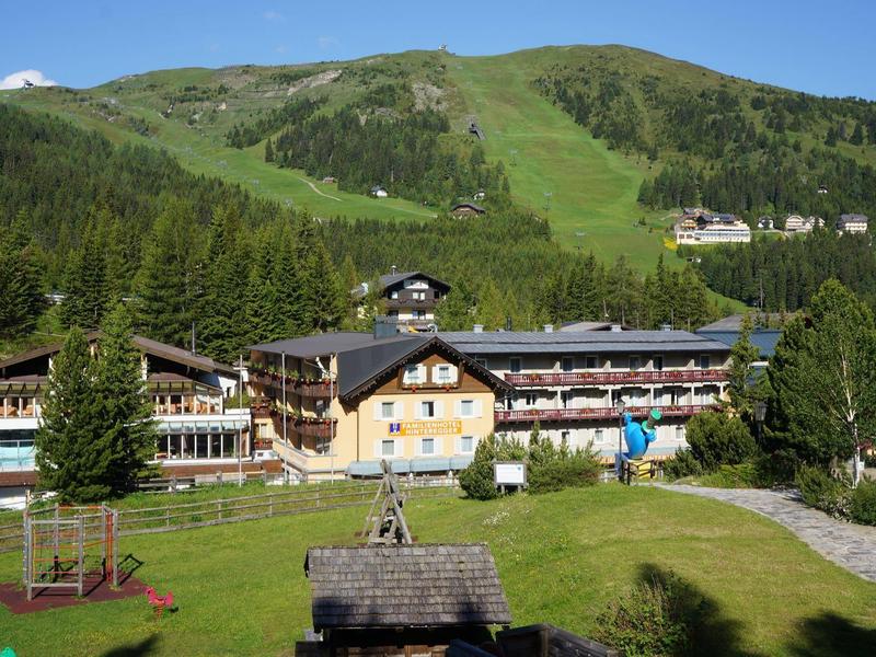 Alpenhotel vor grünem Berg mit Wiesen, Bäumen und Spielplatz im sonnigen Tal.