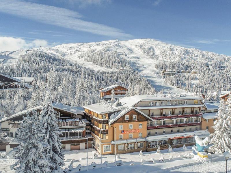 Großes Holzhaus umgeben von schneebedeckten Tannen und Bergen unter blauem Himmel.