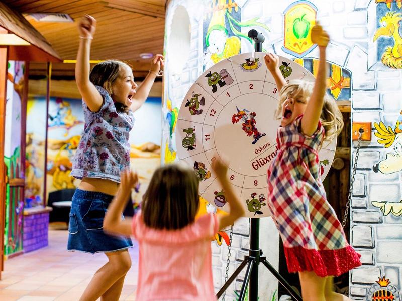 Drei Kinder spielen lebhaft vor einem farbenfrohen Glücksrad in einem belebten Spielraum.