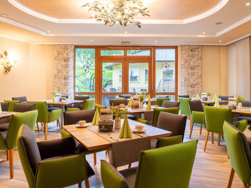 Hell eingerichteter Gastraum mit grünen Stühlen, Holztischen und großen Fenstern.