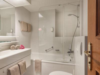 Moderner weißer Badezimmerbereich mit Badewanne, Duschwand, Waschbecken und Toilette, Beleuchtung oben.