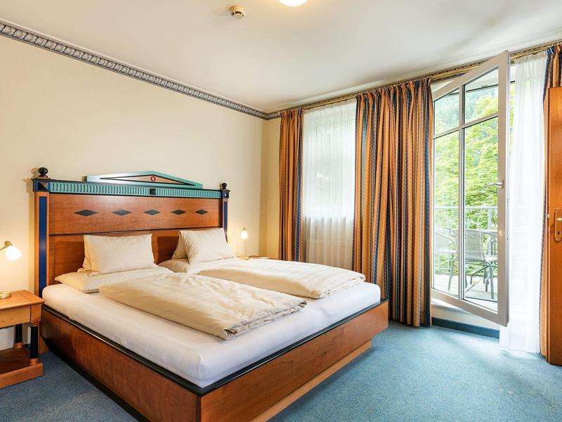 Ein helles Schlafzimmer mit großem Holzbett, zwei weißen Decken, braunen Vorhängen und blauem Teppich.