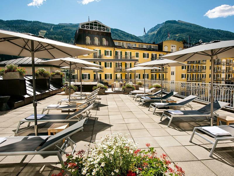 Große Terrasse mit Liegestühlen und Sonnenschirmen, im Hintergrund gelbes Gebäude und Berge.