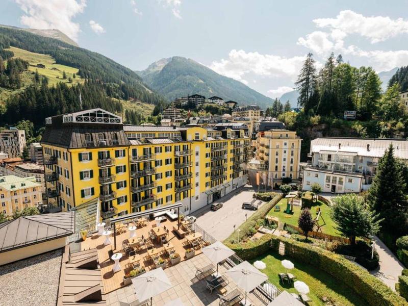 Ein gelbes Apartmentgebäude in grüner Berglandschaft mit Terrasse und Garten unter blauem Himmel.