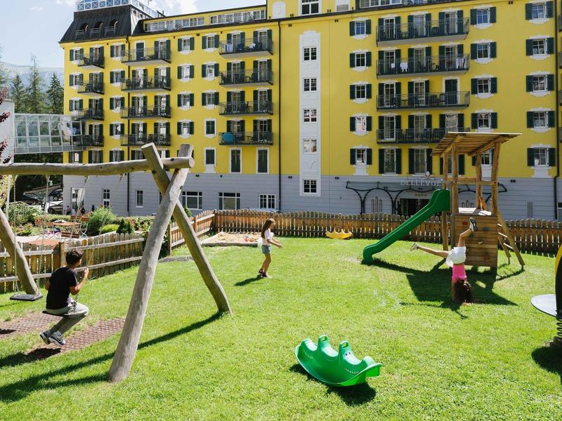 Kinder spielen auf grüner Wiese mit Schaukel und Rutsche vor gelbem Mehrfamilienhaus.