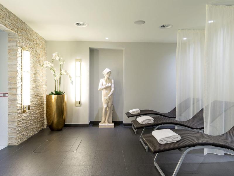 Heller Wellnessraum mit drei Liegen, weißen Handtüchern, Statue und Pflanzen in großem Goldtopf.