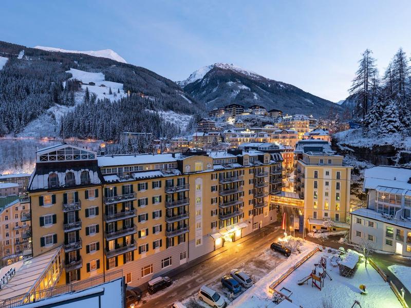 Beleuchtetes Hotelgebäude in verschneiter Berglandschaft bei Dämmerung mit Skihang im Hintergrund.