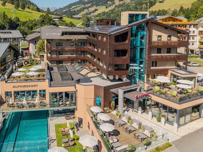 Modernes Hotel mit mehreren Stockwerken, Terrasse mit Sitzbereichen und Pool vor grünen Bergen.