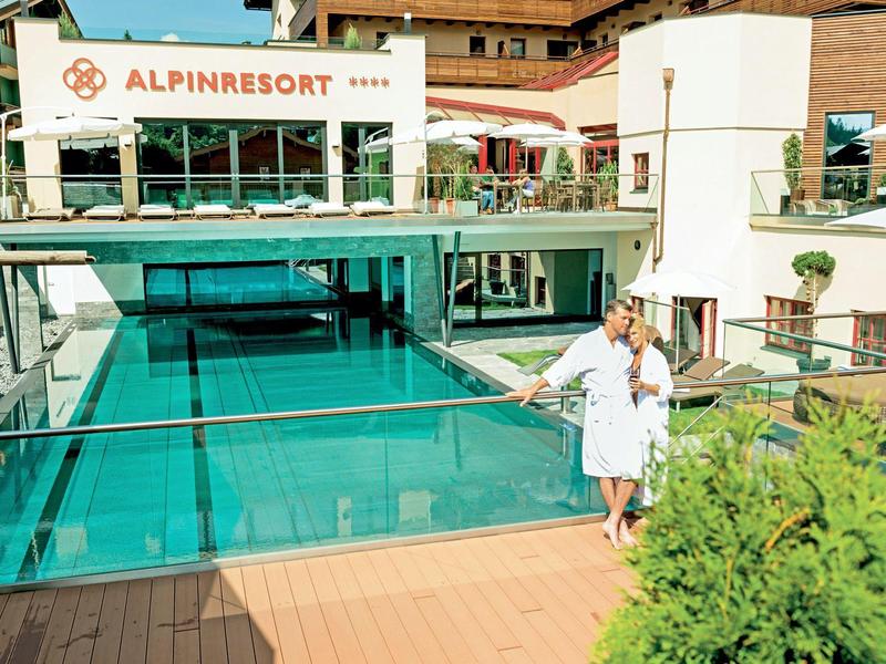 Basen na zewnątrz ze szklanymi ścianami i budynkiem hotelu w tle, dwie osoby w szlafrokach przy basenie