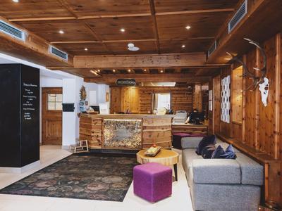 Acogedor vestíbulo de hotel con paredes de madera, alfombra, sofá gris y pufs coloridos.