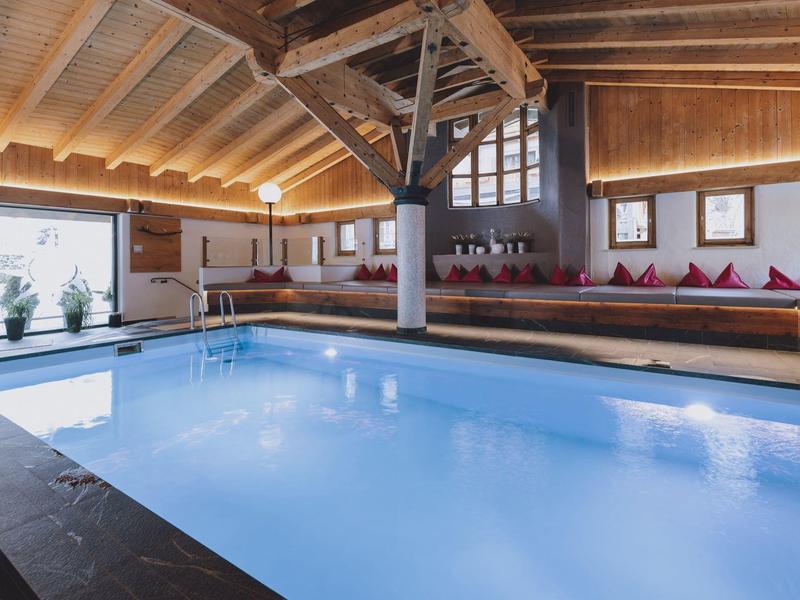Vnitřní bazén s dřevěným stropem, posezením s červenými polštáři a velkými okny pro přirozené světlo.
