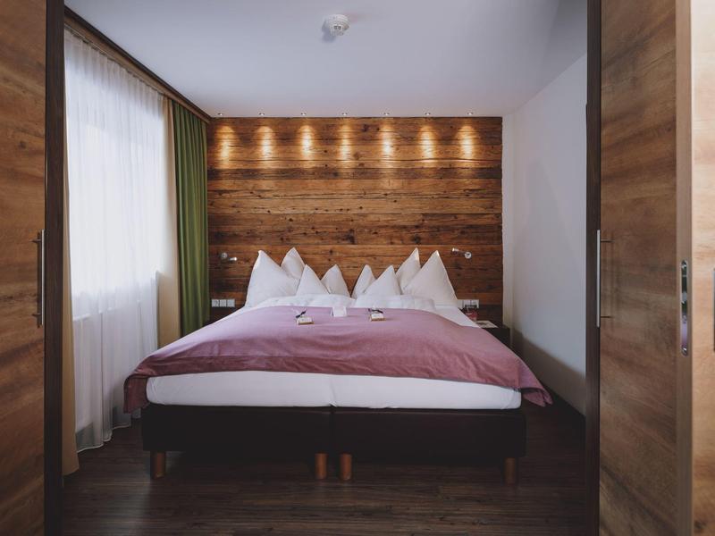 Moderní ložnice s manželskou postelí, dřevěnou stěnou a nepřímým osvětlením.