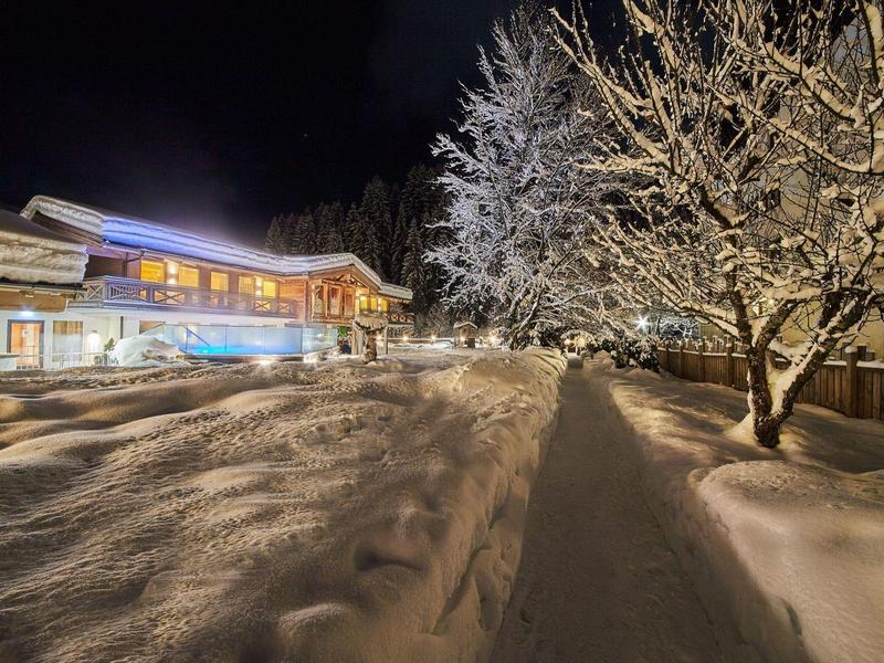 Sněhem pokrytá cesta v noci vede k osvětlené budově s zasněženými stromy vpravo.