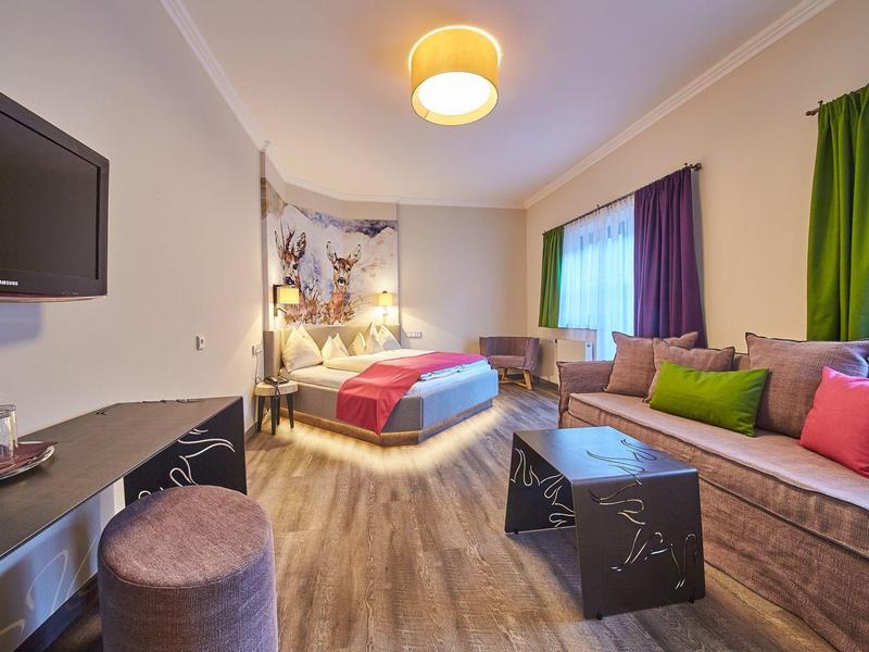 Moderní hotelový pokoj s manželskou postelí, pohovkou, televizí s plochou obrazovkou a dřevěnou podlahou.