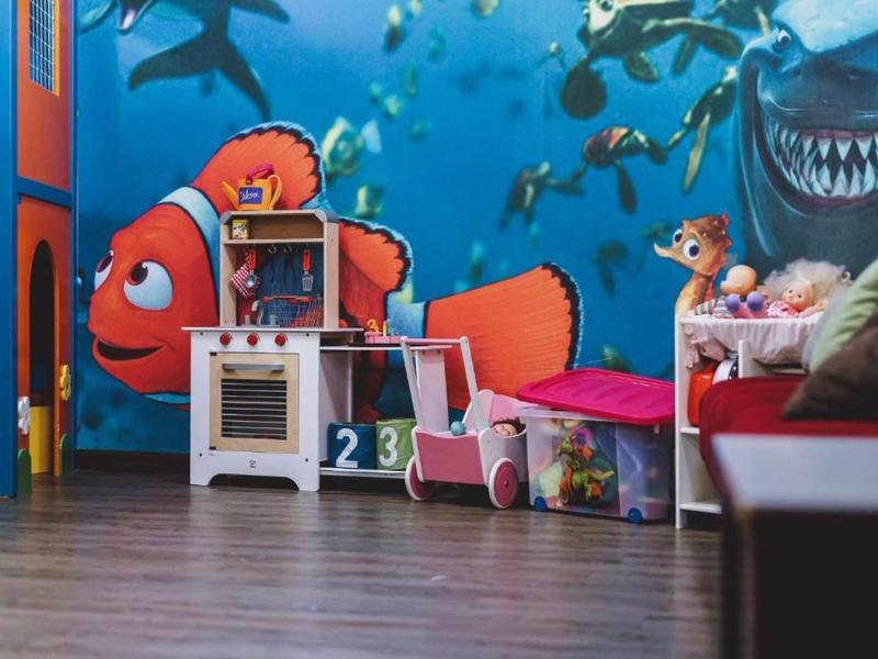 Barevný dětský pokoj s rybí malbou, dětskou kuchyňkou a autíčkem na dřevěné podlaze.