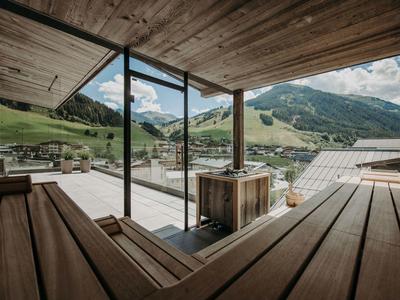 Blick von einer Holzterrasse mit Heißwasserbad auf grüne Hügel und Berge unter bewölktem Himmel.
