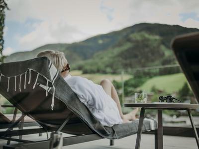Person liegt entspannt auf Liegestuhl mit Bergblick, vor ihr steht ein Tisch mit Getränk.