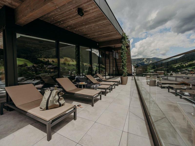 Moderne Terrasse mit braunen Liegen, Glasgeländer und Berglandschaft im Hintergrund.