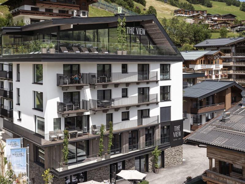 Moderner Hotelbau mit Glasbalkonen und Dachterrasse in alpiner Berglandschaft mit umliegenden Holzhäusern.