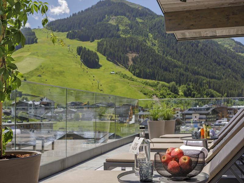 Berglandschaft mit grünen Hügeln, Terrasse mit Liegestühlen, Glasgeländer und Obstschale im Vordergrund.