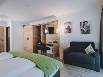 Camera d'hotel luminosa con due letti, copriletto verde, TV e poltrona vicino alla finestra.