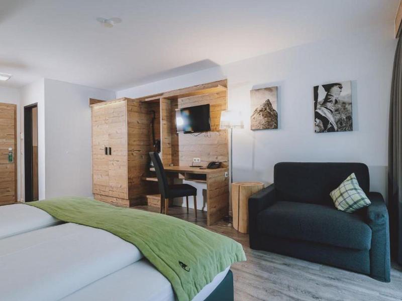 Camera d'hotel luminosa con due letti, copriletto verde, TV e poltrona vicino alla finestra.